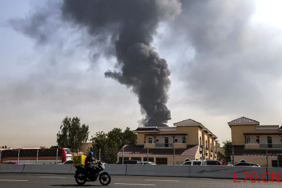 一栋建筑后方升起浓烟，前景是一条高速公路，一辆摩托车正疾驰而过。这是上月遇袭后，迪拜国际机场升起浓烟的场景。